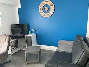 Un salon moderne avec un mur d’accent bleu, un téléviseur à écran plat, une petite table ornée d’un oiseau décoratif, un canapé gris et une bibliothèque.