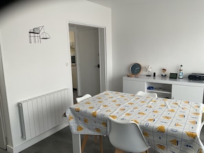 Une pièce avec une table recouverte d’une nappe à motifs, un radiateur blanc, un bureau blanc avec une horloge, une radio et une petite figurine.