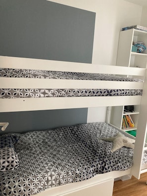 Een stapelbed met beddengoed met een patroon, een wit wandrek met boeken, en een grijze muur.