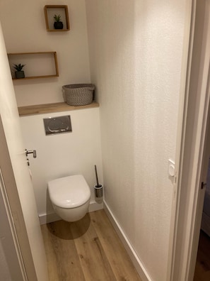 Een kleine badkamer met een toilet, een plankje met een plant en een mand, en een wc-papierrolhouder aan de muur.