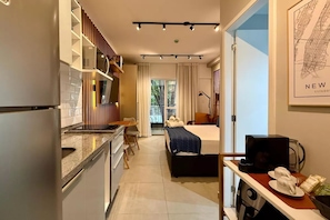Habitación de hotel compacta con kitchenette, una cama con manta azul, un escritorio con computadora y vista al exterior.