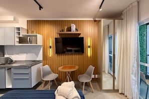 Habitación de hotel moderna con kitchenette, televisor de pantalla plana montado en una pared de madera, una pequeña mesa redonda con dos sillas y un amplio ventanal con cortinas.