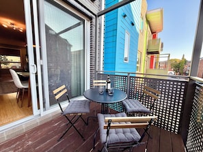 A balcony with a table and chairs, a bottle of wine, and a view of colorful buildings. - Walkable downtown Dayton MCM townhome with garage and balcony! (Dayton)