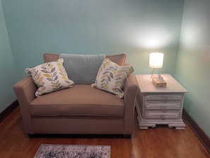 Un canapé marron agrémenté de coussins à motifs de feuilles, une table de chevet blanche avec une lampe, et un tapis à motifs posé sur un plancher en bois.