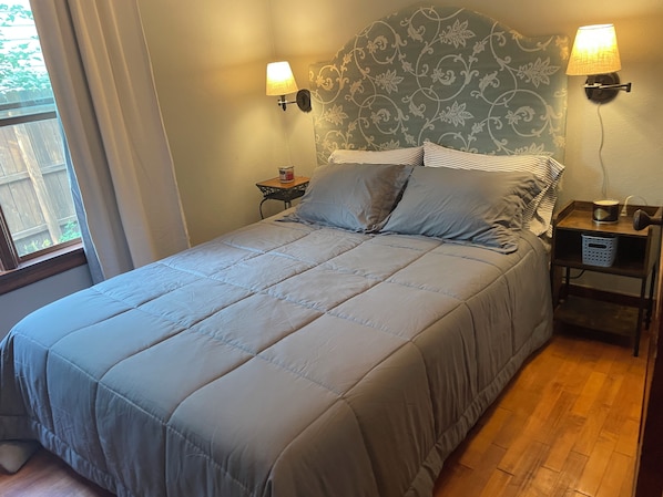 Un lit avec une tête de lit, deux lampes de chevet, une table de chevet avec une lampe et un panier, et une fenêtre avec des rideaux.