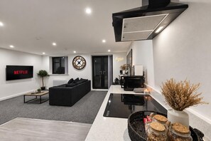 En moderne stue med en sort sofa, et fladskærms-tv, et vægur og et tekøkken med mikrobølgeovn og kaffemaskine.