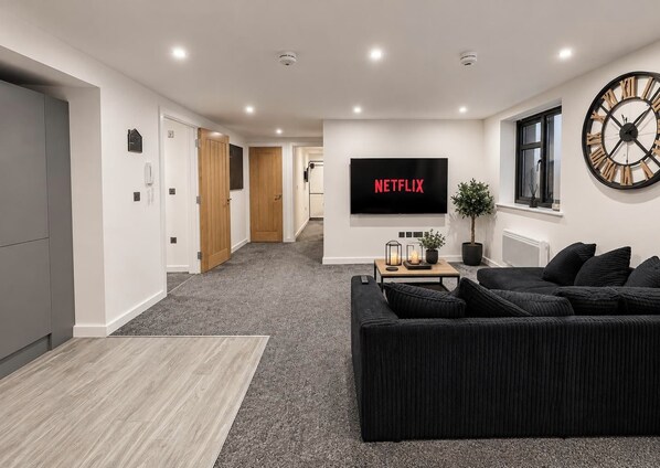 En moderne stue med et stort fladskærms-tv, der viser 'NETFLIX', en sort sofa, et sofabord og en potteplante.