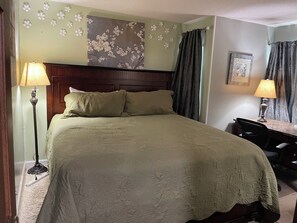 Una cama bien hecha con cabecera, dos lámparas de noche, un escritorio con silla y una pared con estampado floral.