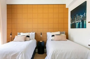 A hotel room with two beds, a headboard with a geometric pattern, a nightstand with a lamp, and a cityscape painting on the wall. - Good Vibe Rooms (Bilbao)