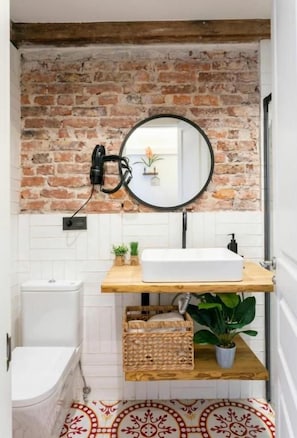 A bathroom with a brick wall, a round mirror, a white sink, and a wooden shelf with a plant. - Good Vibe Rooms (Bilbao)