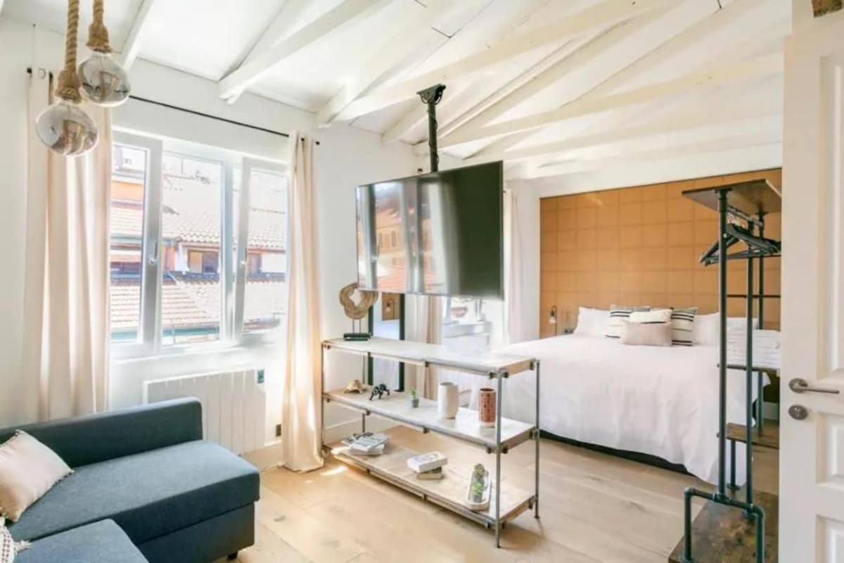 A hotel room with a bed, a sofa, a TV on a stand, a shelf with items, and a view of buildings outside.