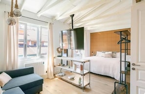 A hotel room with a bed, a sofa, a TV on a stand, a shelf with items, and a view of buildings outside. - Good Vibe Rooms (Bilbao)