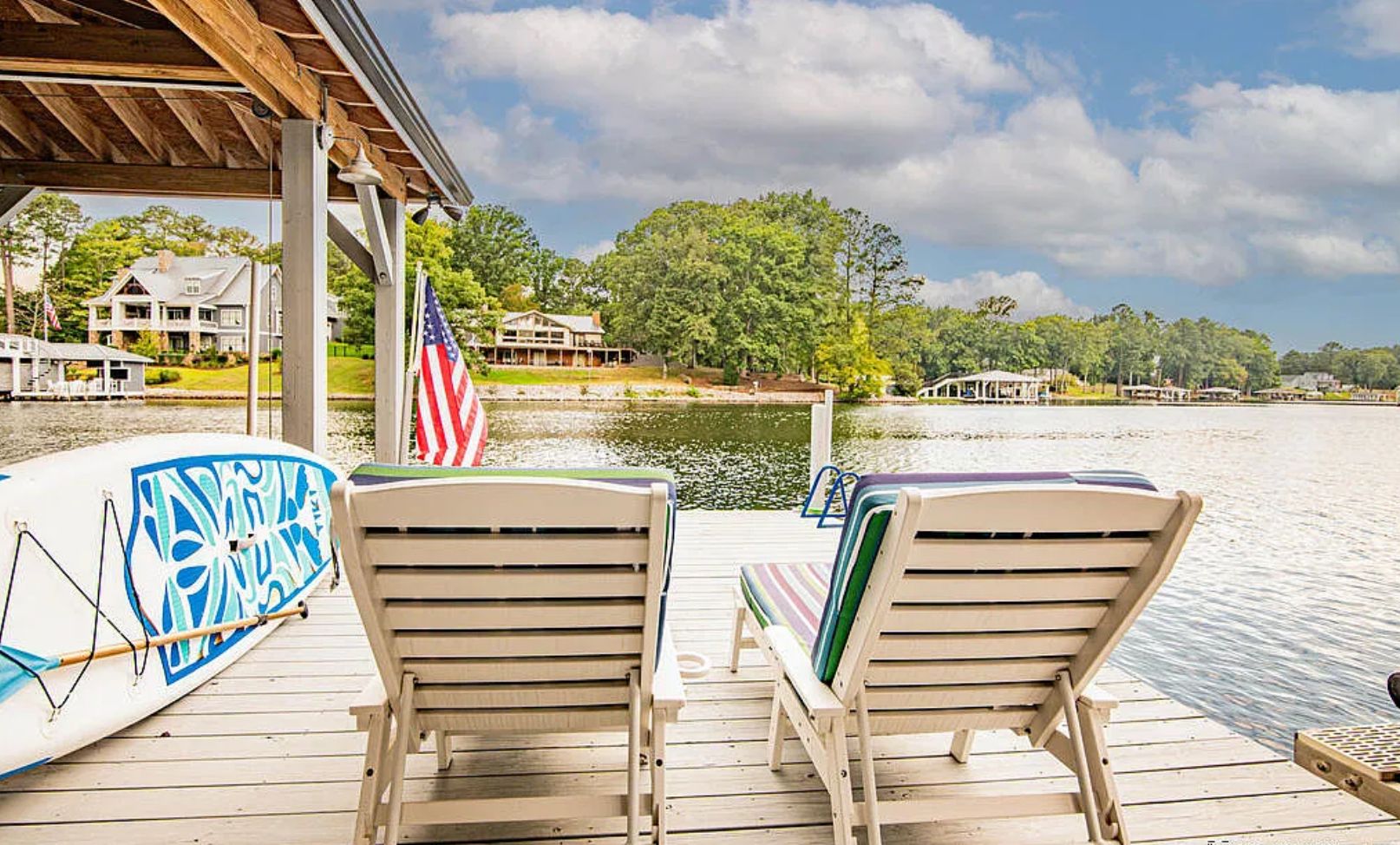 A wooden deck with two lounge chairs and a surfboard, overlooking a body of water with a dock and houses in the background.