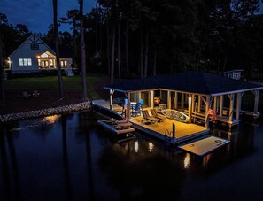 A house with a dock extending into a body of water, a covered seating area with chairs and a table, and a flagpole with an American flag.