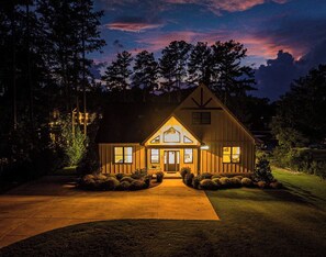 A two-story wooden house with a front porch, illuminated windows, and a well-maintained lawn.