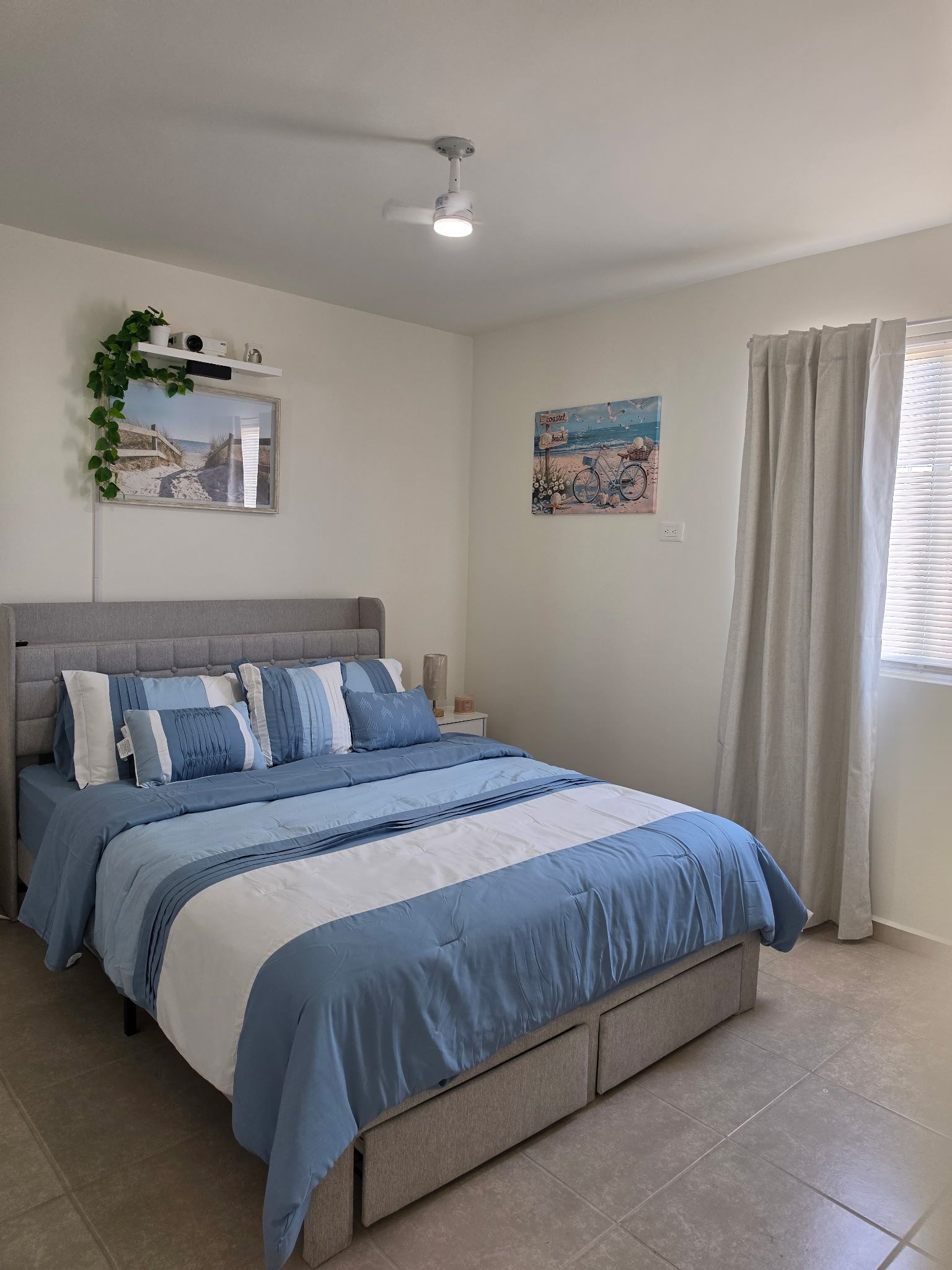 Un dormitorio con cama, ventilador de techo, repisa con planta, cuadro y ventana con cortinas.