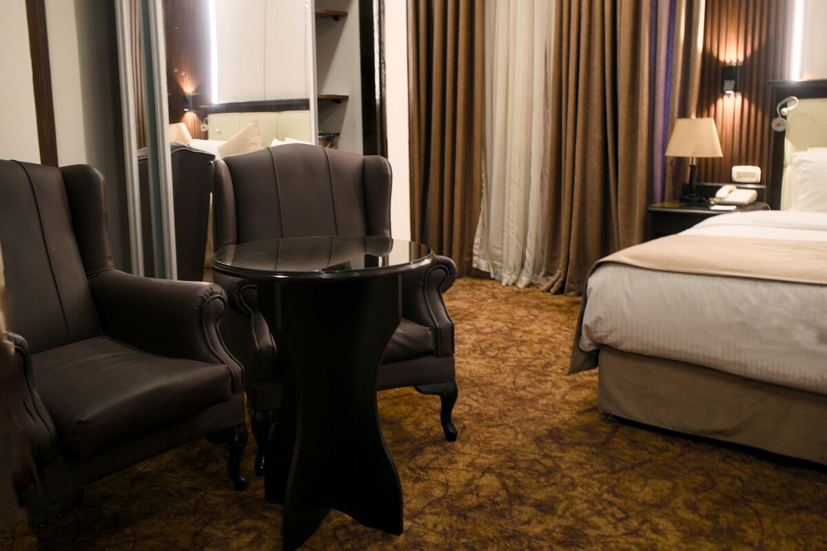 A hotel room with a bed, two armchairs, a small round table, a nightstand with a lamp, and a view of a cityscape through large windows.
