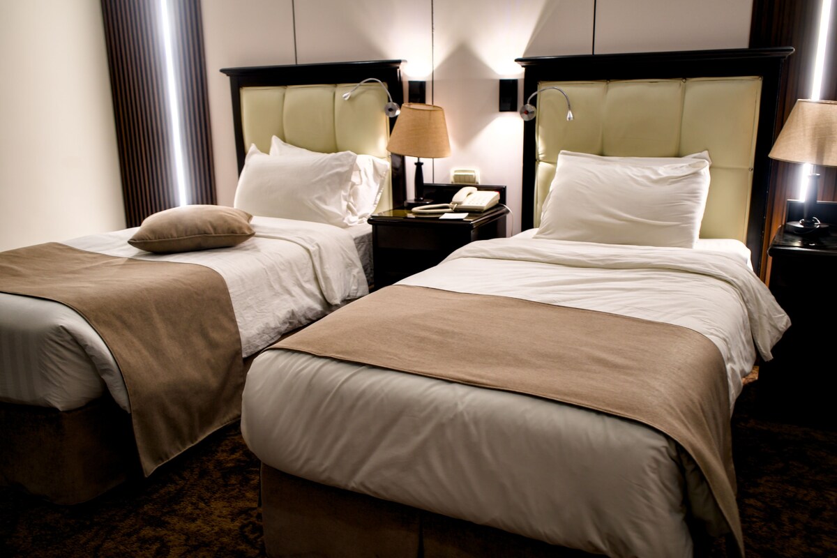 A hotel room with two beds, each with a headboard, a bedside table with a lamp, and a telephone.