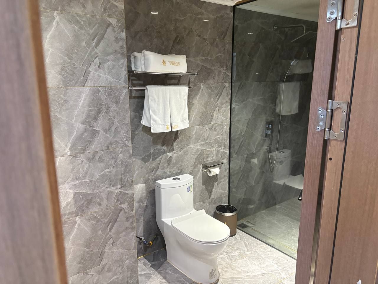 Une salle de bain moderne avec un mur en marbre, des toilettes, une douche en verre et un porte-serviettes avec des serviettes pliées.