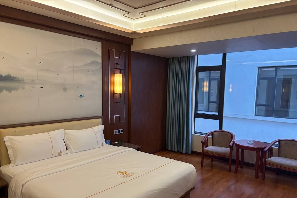 A hotel room with a large bed, a desk with two chairs, a window with curtains, and a wall with a decorative panel.