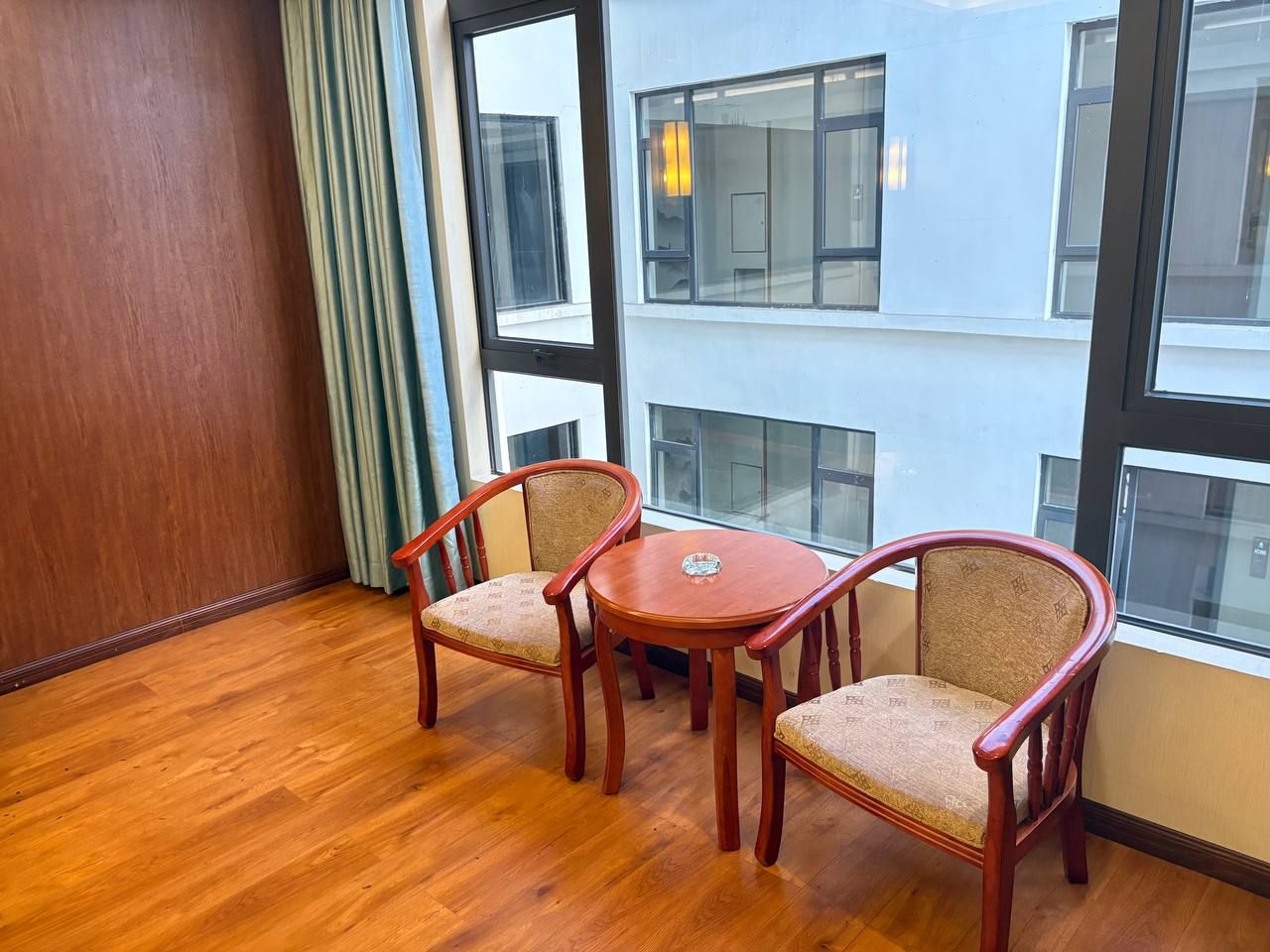 Une pièce dotée d’un plancher en bois, de deux chaises en bois avec des sièges rembourrés, d’une petite table ronde et de grandes fenêtres donnant sur un immeuble.