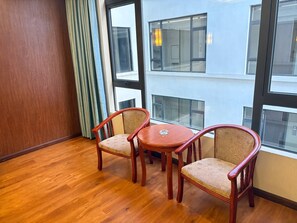 Una habitación con piso de madera, dos sillas de madera con asientos acolchados, una mesita redonda y ventanas amplias que ofrecen vistas a un edificio.