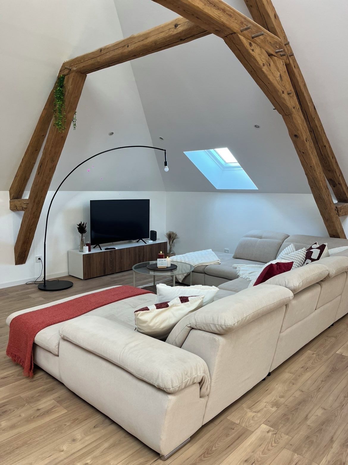 A modern loft-style living room with a high ceiling, a large window, a sofa, a TV, and a wooden beam structure.