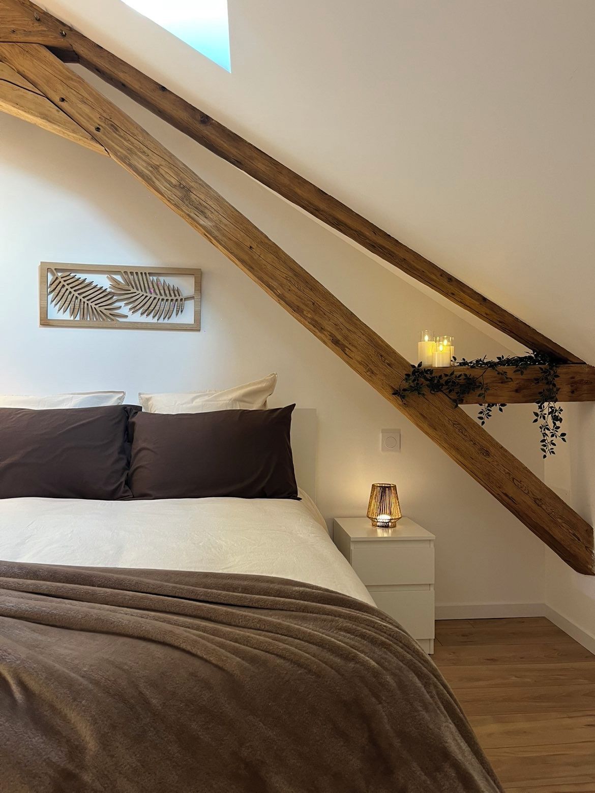 A bedroom with a sloped ceiling, a bed with dark bedding, a bedside table with a lamp, and a wall-mounted decorative piece.