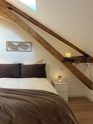 A bedroom with a sloped ceiling, a bed with dark bedding, a bedside table with a lamp, and a wall-mounted decorative piece. - New Listing (Sains-du-Nord)
