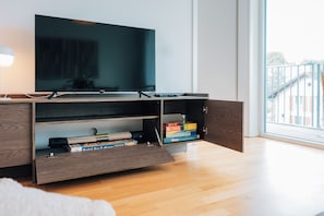Een moderne woonkamer met een flatscreen televisie bevestigd aan een houten kast, een verschuifbare plank met gezelschapsspellen, en een groot raam met uitzicht op buiten.
