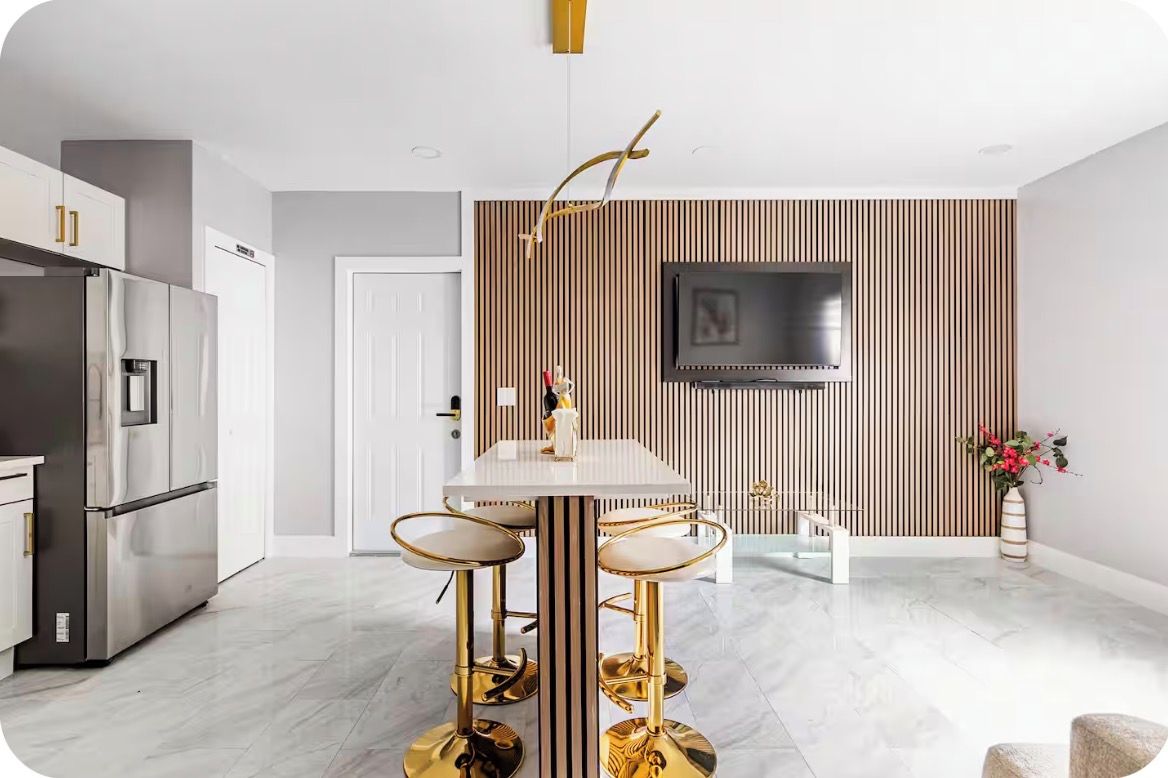 A modern kitchen with a built-in refrigerator, a dining table with bar stools, and a flat-screen TV mounted on a wooden panel wall.