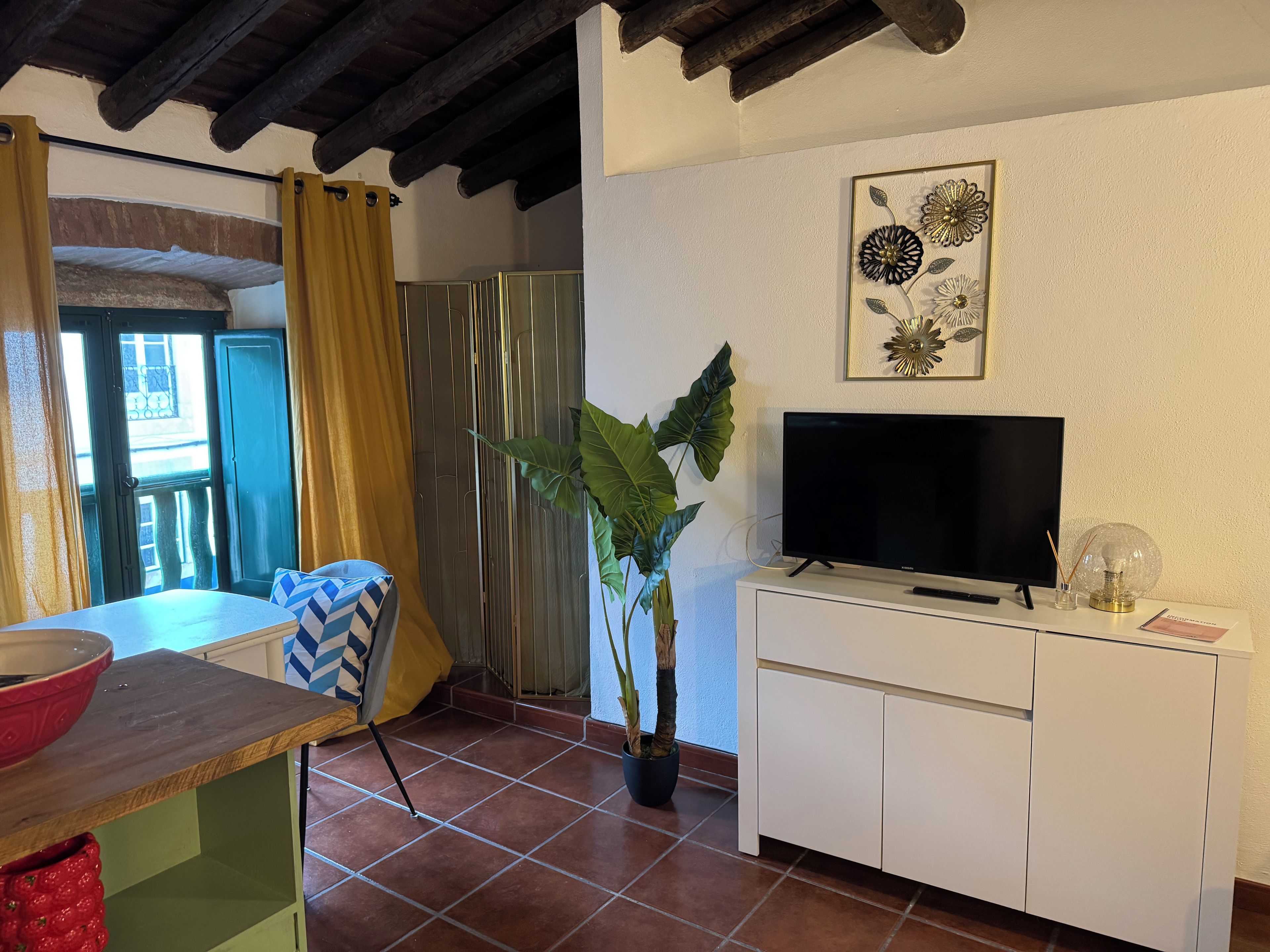 Ein Zimmer mit Fernseher auf einer weißen Kommode, einer Topfpflanze, einem Fenster mit Vorhängen und einer Tür mit Blick nach draußen.