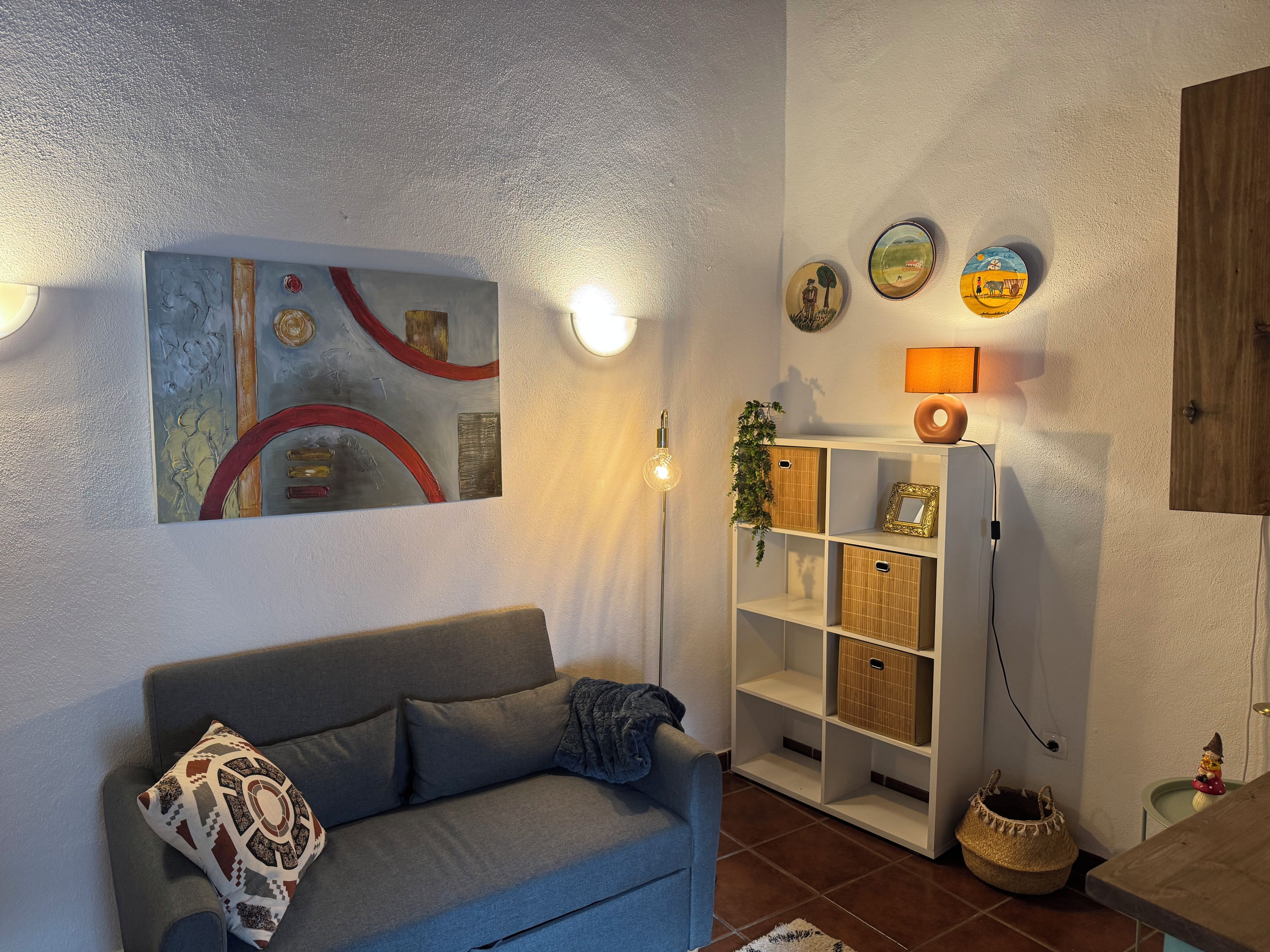Una sala de estar con un sofá gris, un cuadro abstracto colgado en la pared, una estantería blanca con cestas y una mesita con una lámpara.