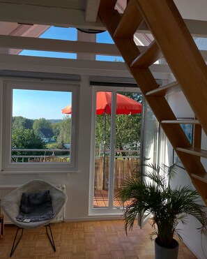 A room with a wooden floor, a chair, a potted plant, and a view of a balcony with a red umbrella. - New Listing (Kürten)