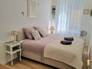Een slaapkamer met een bed, nachtkastje, lamp en een raam met dunne gordijnen.