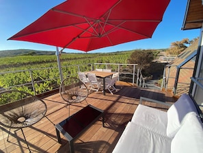 Une terrasse avec une table et des chaises, un parasol rouge et une vue sur un vignoble.