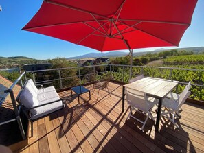 Une terrasse en bois avec une table et des chaises, un parasol rouge et une vue sur les vignes et les collines.
