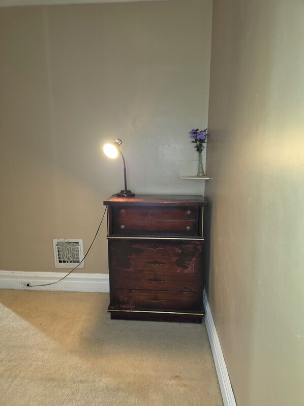 Une chambre d’angle avec une commode en bois, une lampe, une prise de courant et une petite étagère ornée de fleurs.