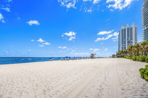 A wide sandy beach with a clear blue sky, palm trees, and tall buildings in the background. - 
26floor PH Marenas Updated Gorgeous Condo w/Direct Beach Access, Washer Dryer (Sunny Isles Beach)