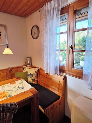 Habitació acollidora amb una banca de fusta, una taula amb estovell floral, un rellotge a la paret i una finestra amb cortines translúcides.