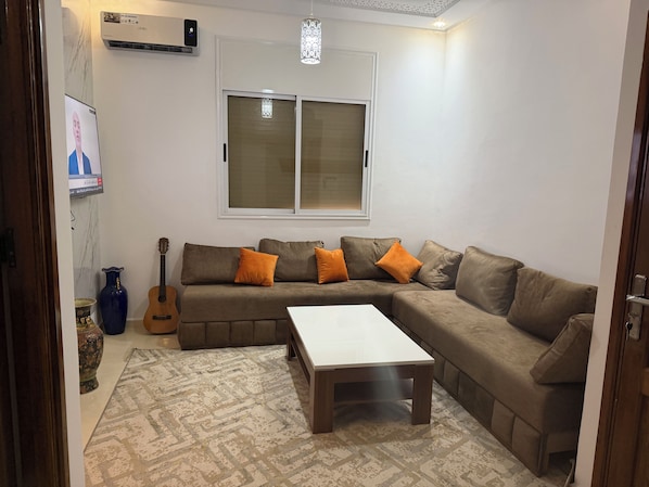 A living room with a sectional sofa, a coffee table, a guitar, and a TV mounted on the wall.