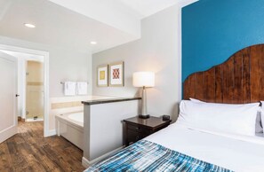 Ein Hotelzimmer mit einem Bett, einem Nachttisch, einer Lampe und einem Badezimmer inklusive Badewanne und Dusche.