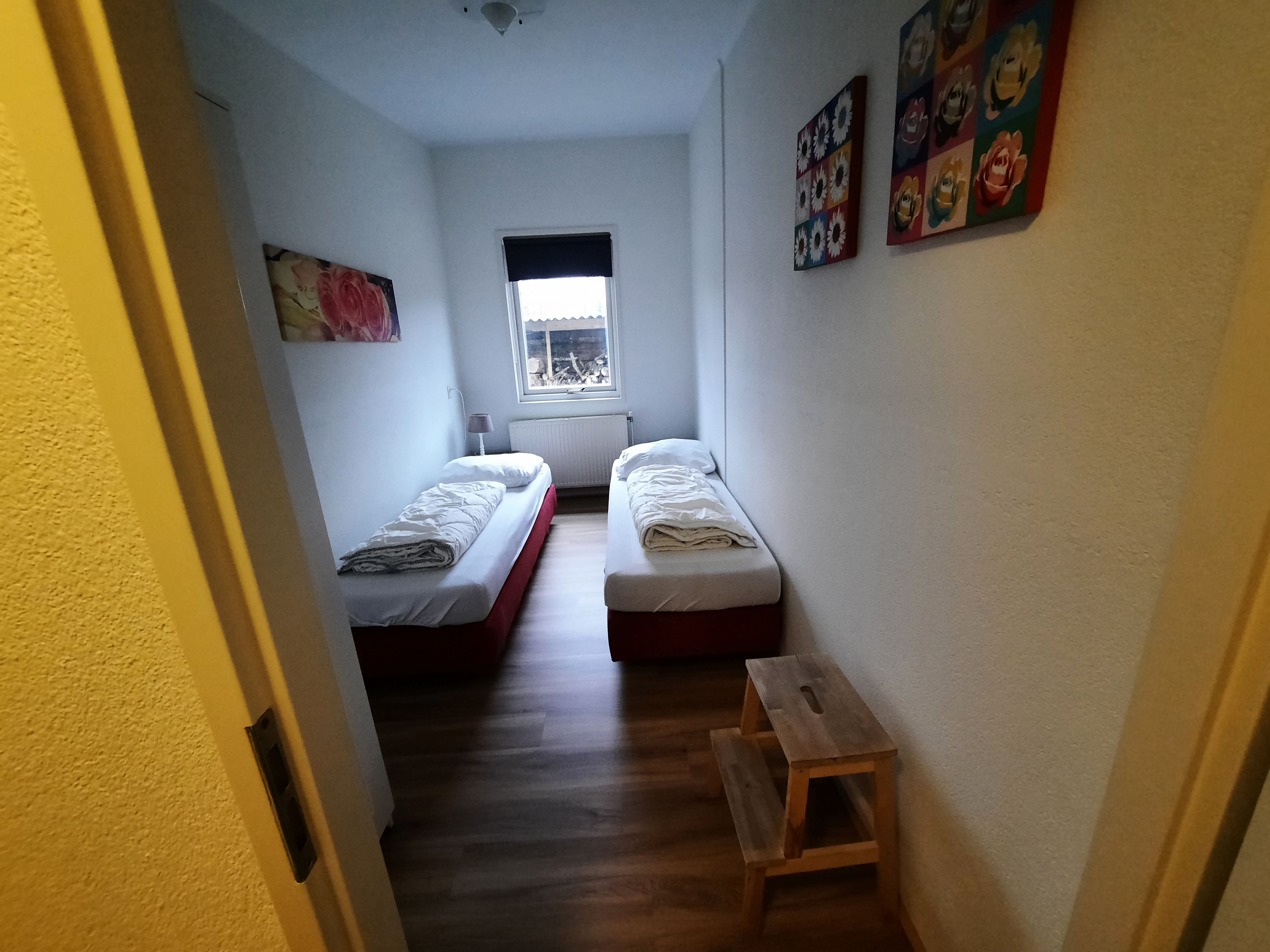 Une petite pièce étroite avec deux lits simples, un petit banc en bois, un radiateur et une fenêtre donnant sur un paysage agréable.