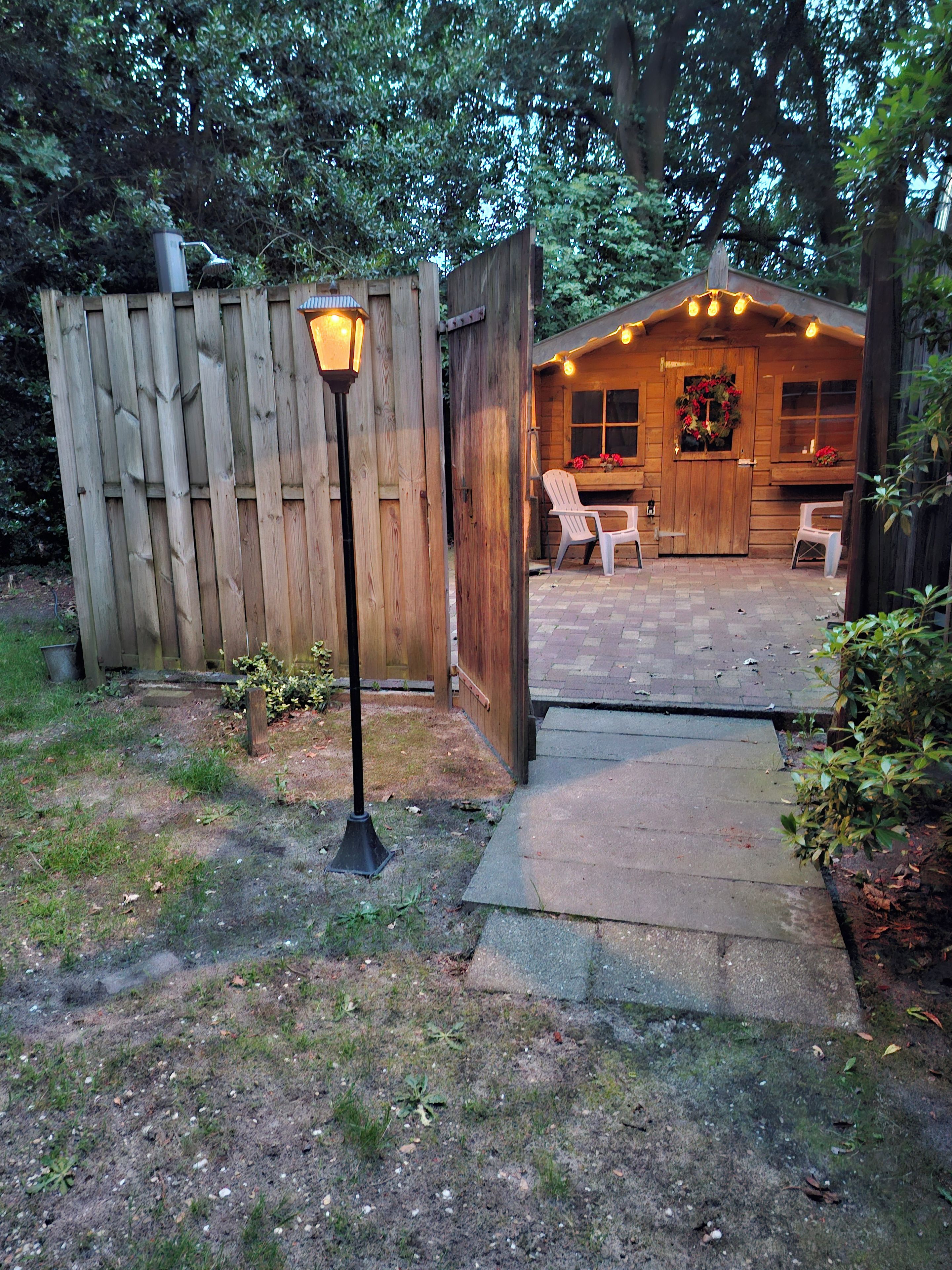 Een houten terras met een pad dat leidt naar een rustieke houten constructie met een raam en een deur versierd met rode bloemen.