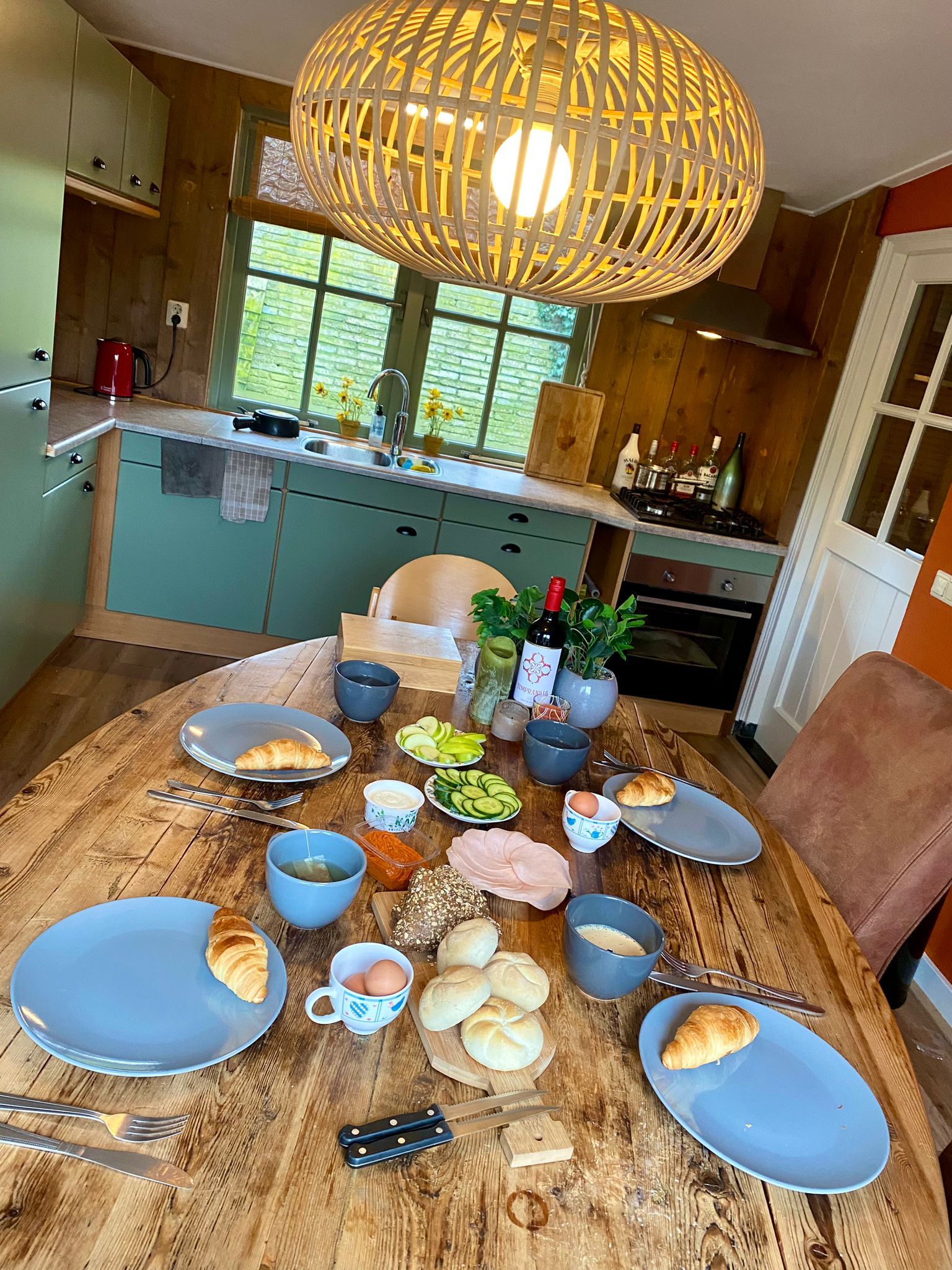 Une table de salle à manger en bois, avec de l’assiette, du pain et des boissons, placée dans une cuisine dotée de meubles en bois et d’un luminaire suspendu.