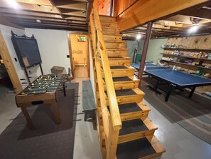 Una escalera de madera conduce a una zona superior con una mesa de futbolín y una mesa de ping pong en una habitación con vigas de madera.