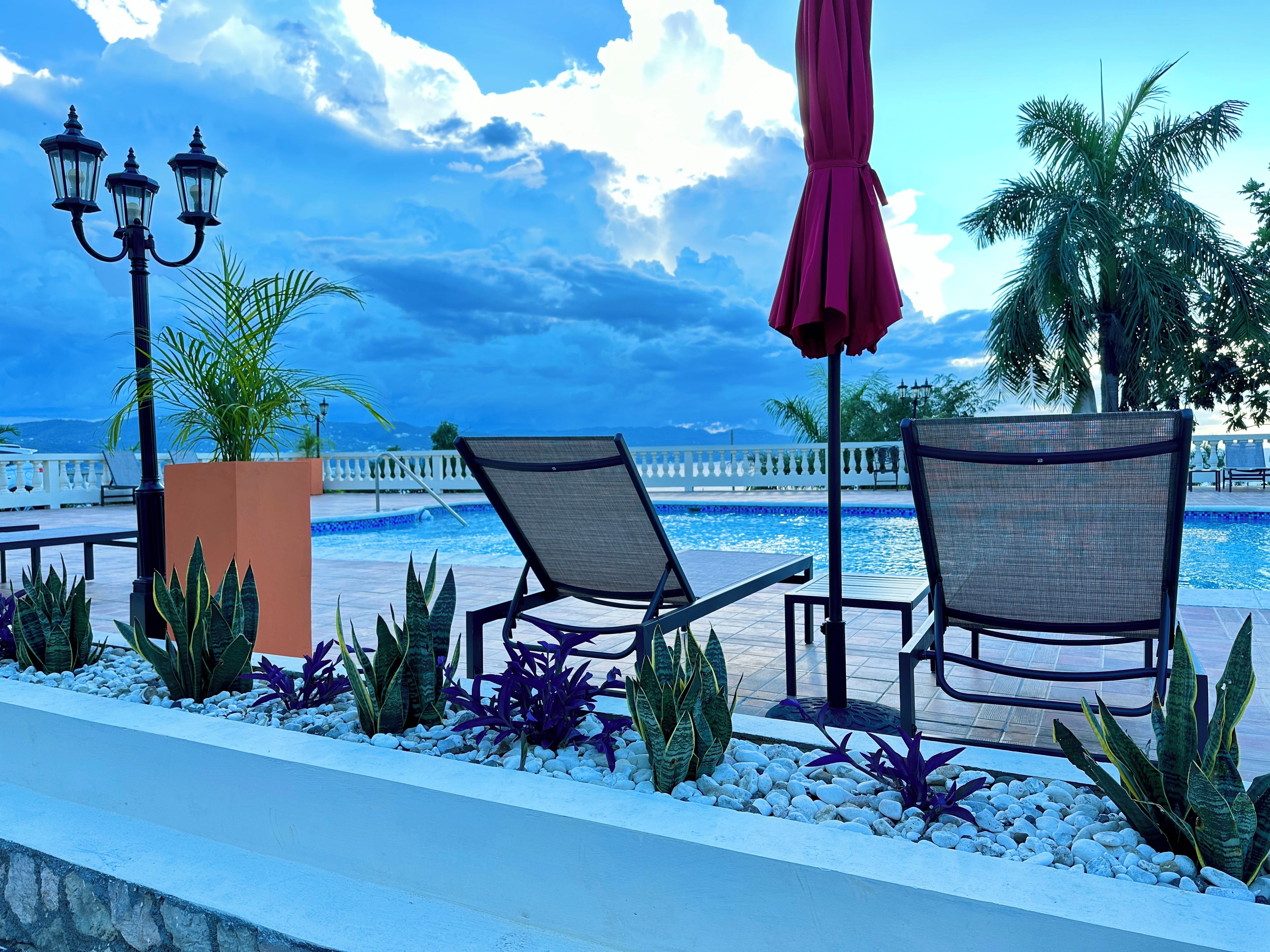 A poolside area with lounge chairs, a lamp post, and a view of the sea.