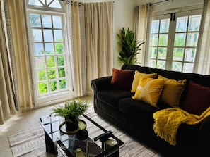 A living room with a black sofa, a glass coffee table with a plant, and a view of greenery through large windows. - Stunning Oceanview 2BR-Pool-Balcony & 24/7 Security (Montego Bay)