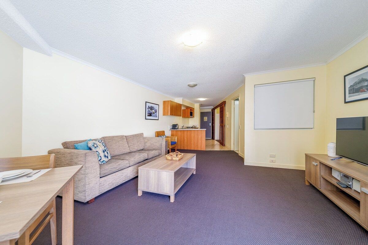 A living room with a sofa, coffee table, TV stand, and a kitchen area in the background.