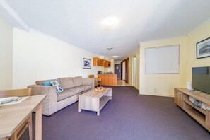 A living room with a sofa, coffee table, TV stand, and a kitchen area in the background. - Mantra Apt - 2nd Floor + Views (Urangan)